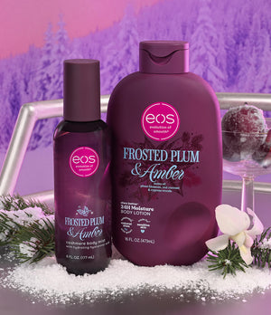 Two EOS body care products in frosted plum and amber packaging, styled with white snow, evergreen branches, and a white orchid against a purple winter forest background.