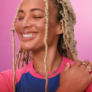 A close-up lifestyle shot of a smiling woman with blonde dreadlocks, wearing a pink and blue colorblock raglan t-shirt and silver jewelry, set against a solid pink background.