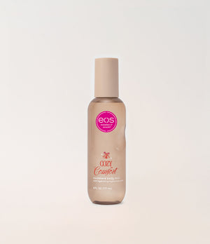 A bottle of EOS Cosmetics Cozy Comfort Body Mist in a frosted beige bottle with a matching beige cap, featuring a pink circular EOS logo on the front, set against a clean off-white background.