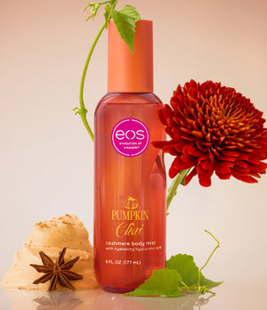 EOS Cosmetics Chui Pumpkin Cashmere Body Mist in a clear bottle showing the orange liquid inside, styled with a red flower, star anise, and ginger on a warm beige background.