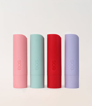Four EOS lip balm tubes in pastel pink, mint green, red, and lavender purple, standing upright in a row against a light off-white background.