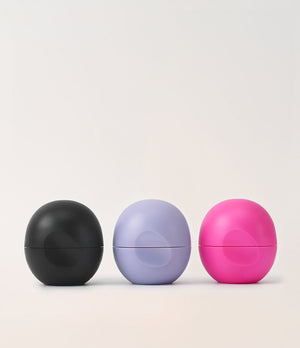 Three matte spherical cosmetic containers in black, light lavender, and bright pink, arranged side by side on a clean off-white background.